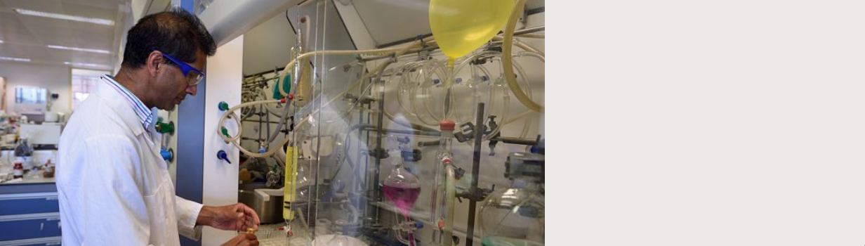 一名男子戴着护目镜，穿着实验服，看着一些含有不同颜色液体的复杂实验室设备。